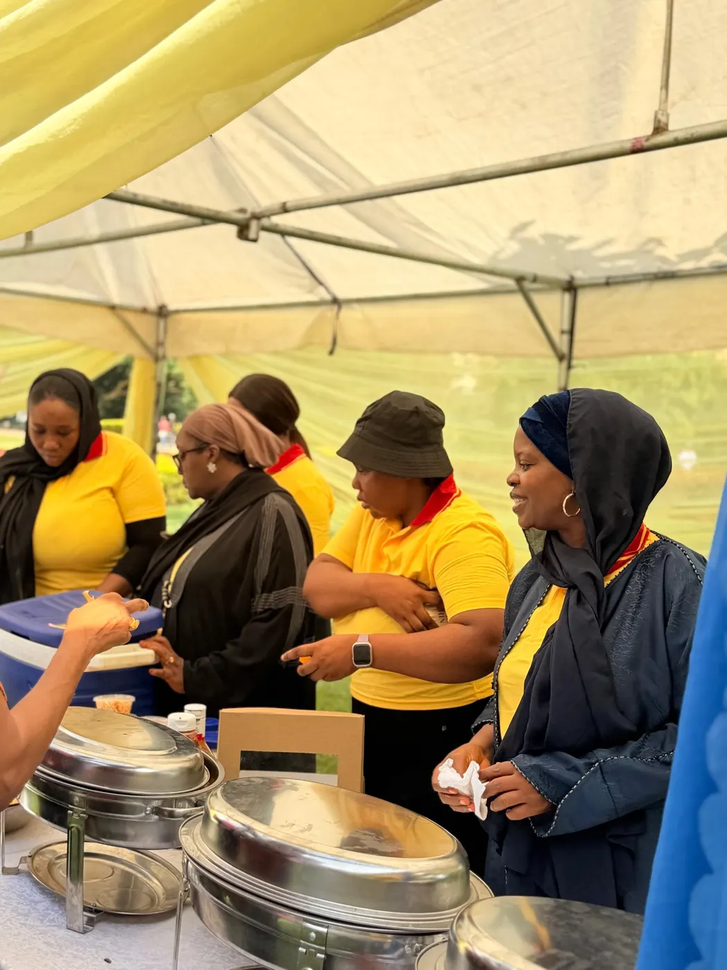 A catering team in yellow shirts serving from chafing dishes under a tent