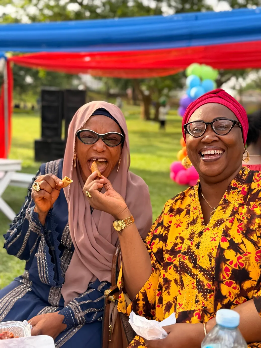 Two women laughing together while tasting small chops outdoors